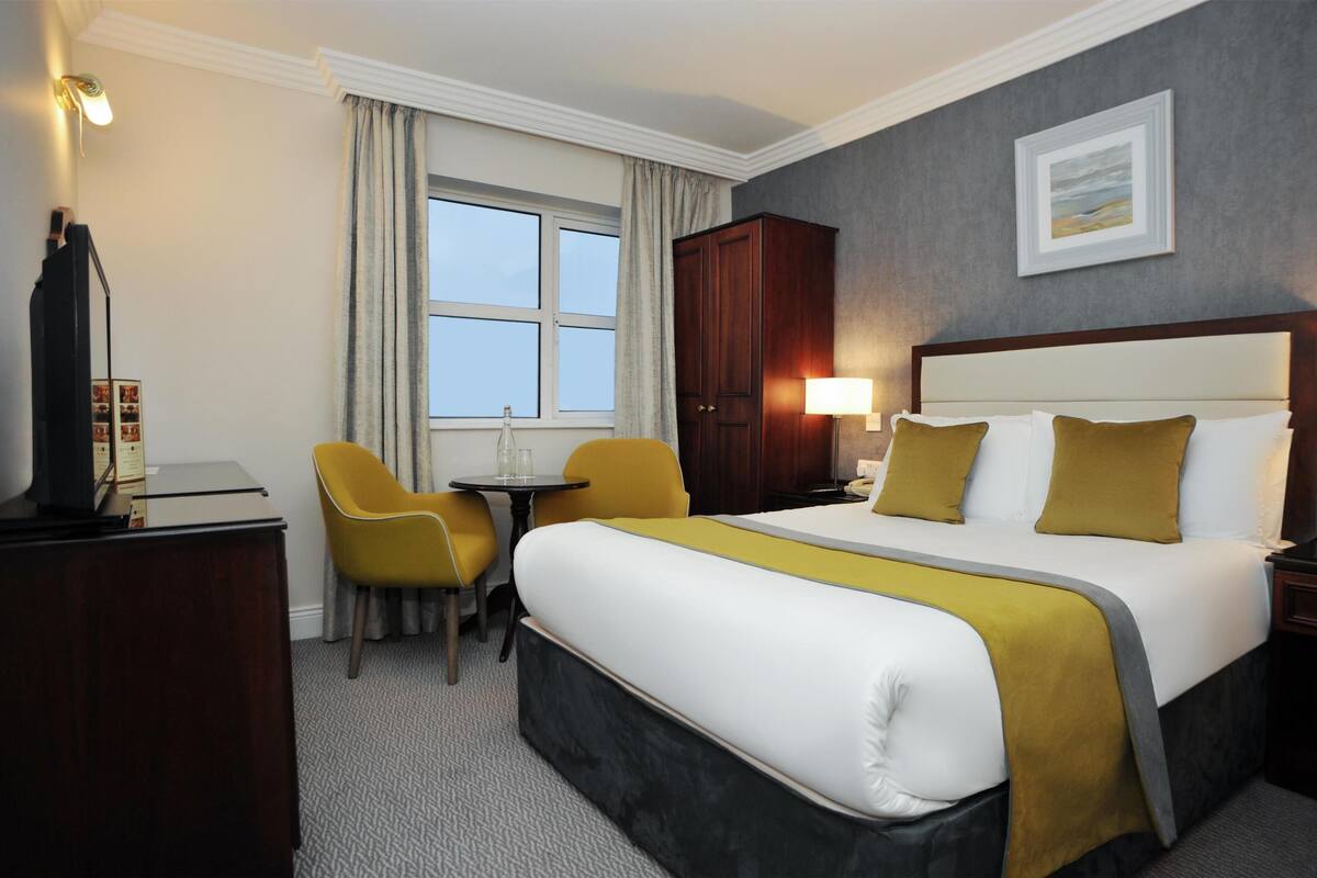 Quarto de hotel com uma cama grande, uma escrivaninha com cadeira, um guarda-roupa e uma janela com cortinas.