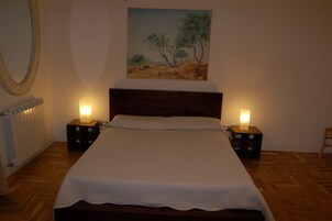Un letto rifatto con un copriletto bianco, due lampade ai comodini, un quadro incorniciato con alberi sulla parete e un pavimento in legno.