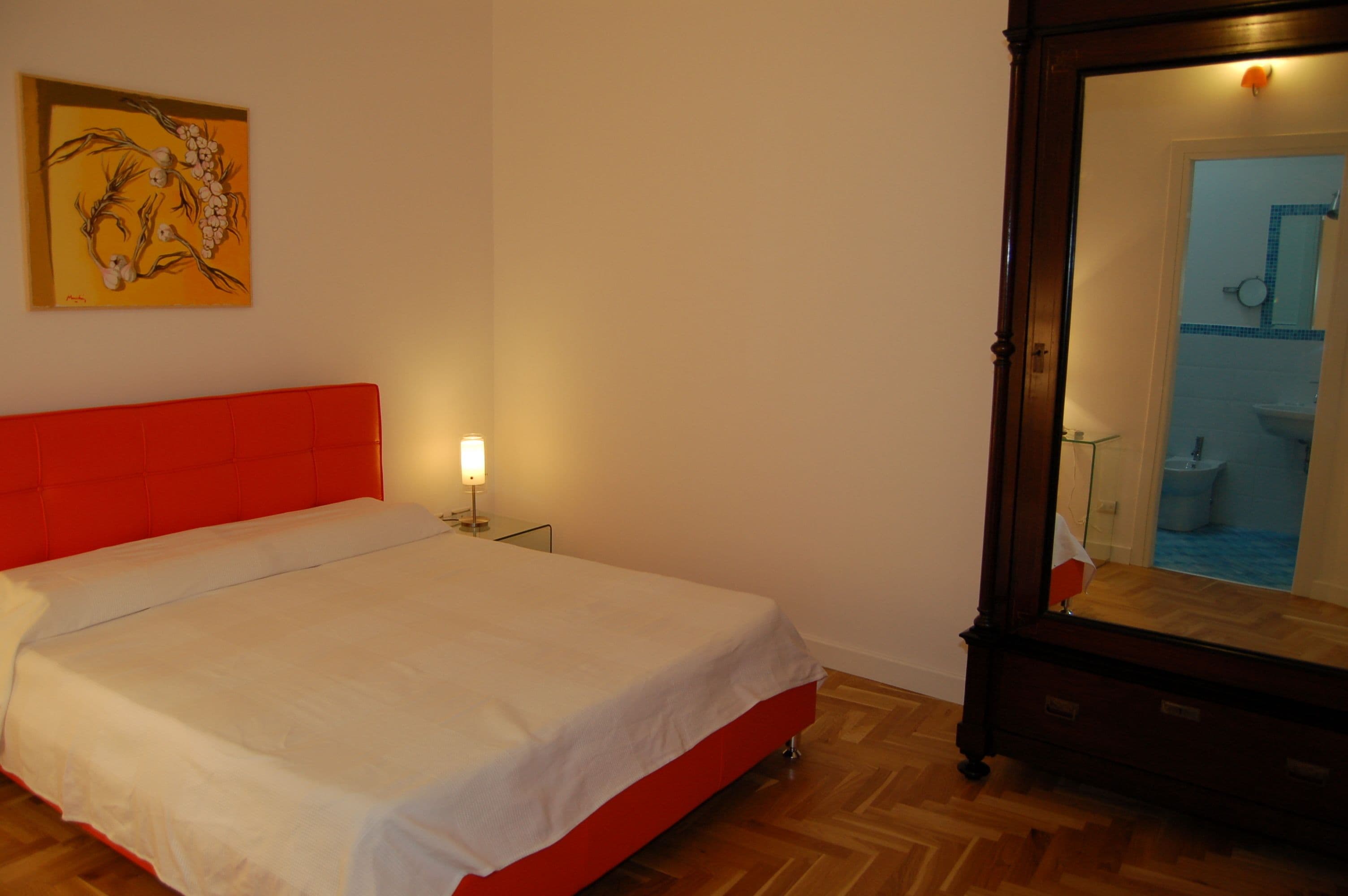 Un dormitorio con un cabecero tapizado en rojo, una colcha blanca, una lámpara de noche, suelo de madera y un espejo que refleja un baño.