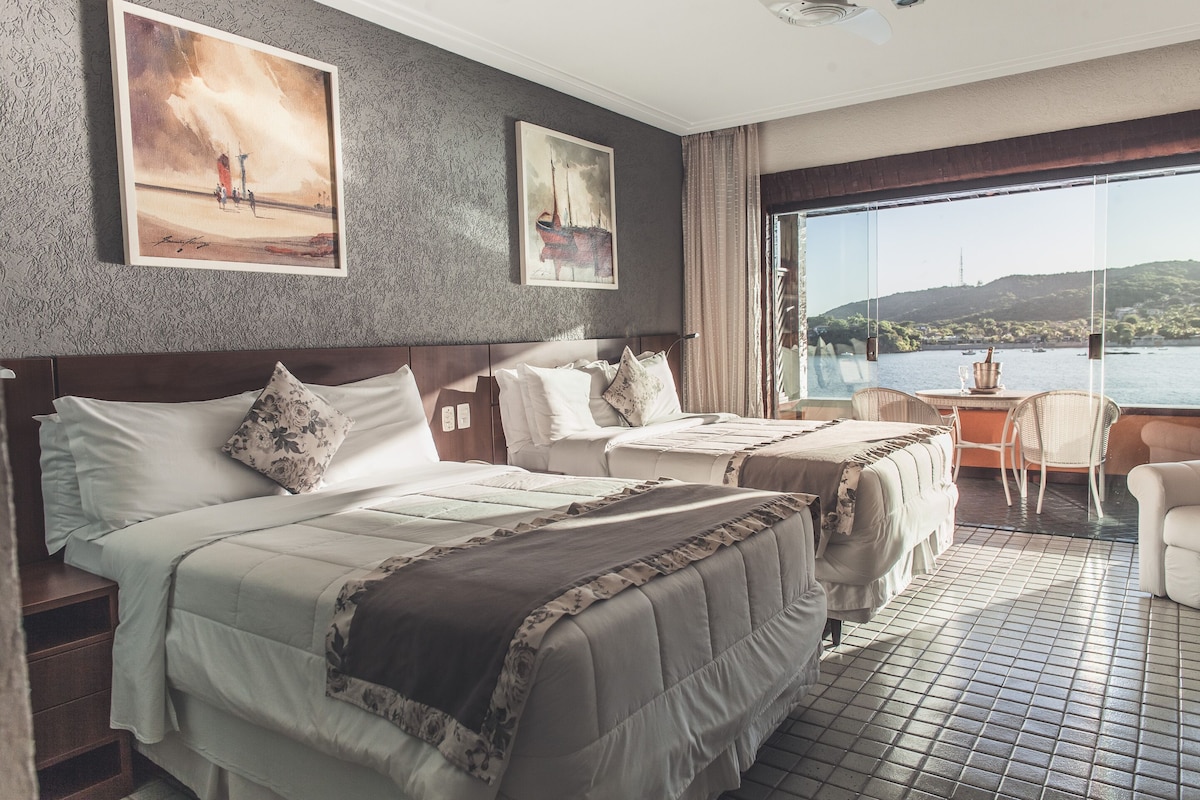 A hotel room with two beds, a large window with a view of a body of water, and artwork on the walls.