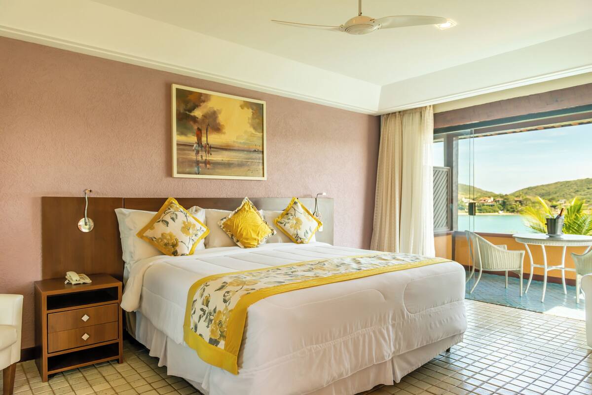 Habitación de hotel con una cama grande, una mesita de noche, un teléfono, un cuadro en la pared y un balcón con vistas al paisaje.