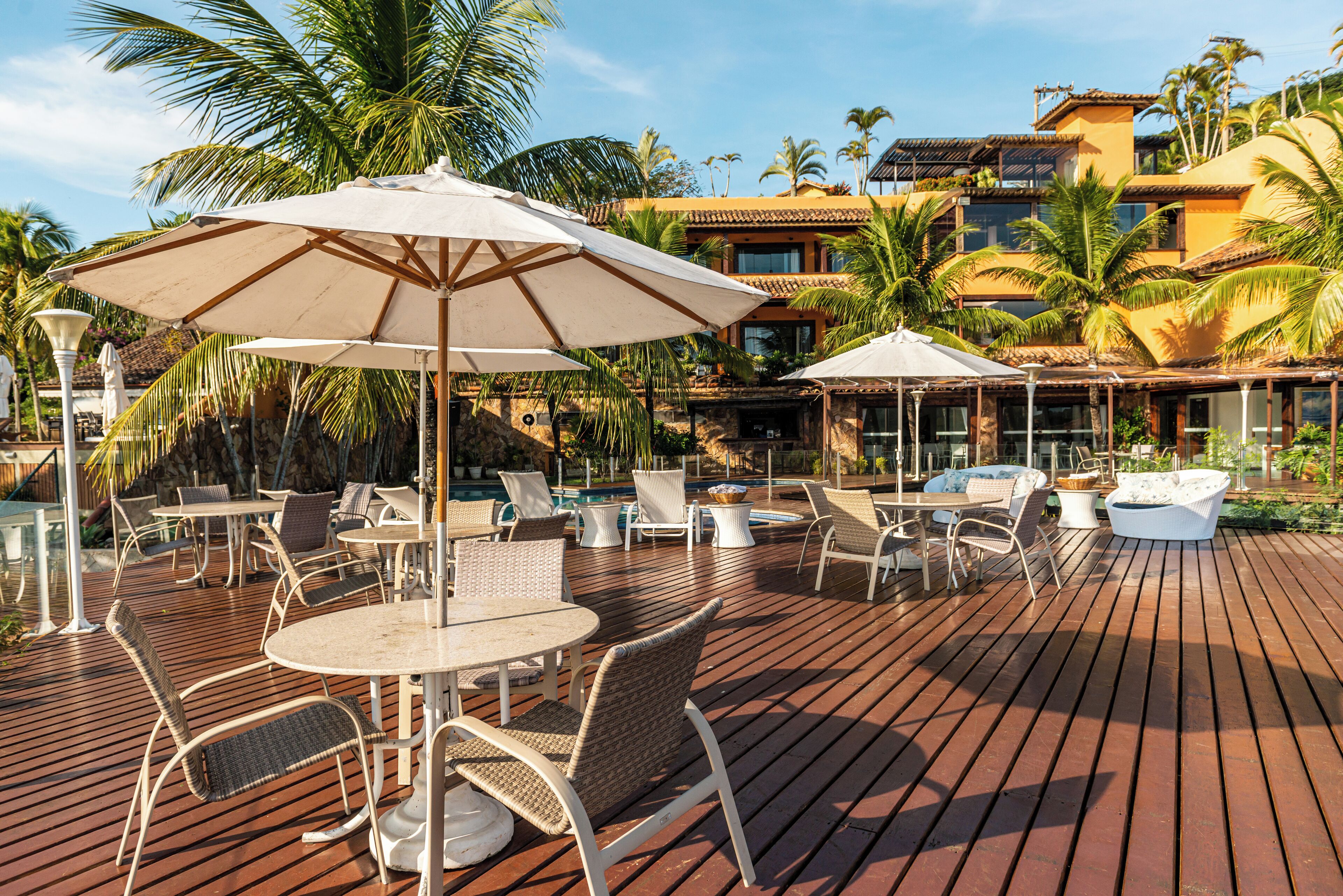 A wooden deck with outdoor seating and umbrellas, surrounded by palm trees and a pool area.