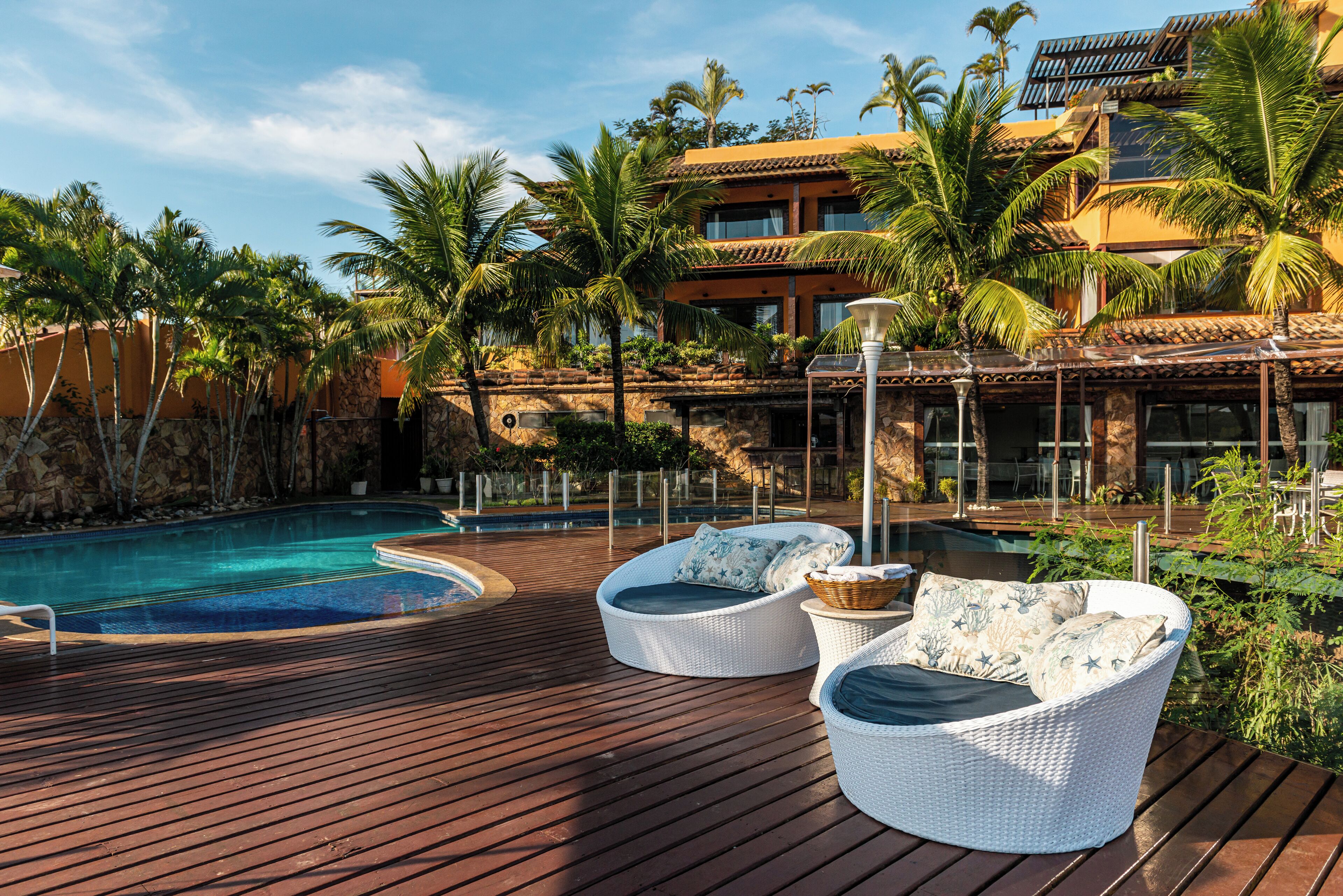 A poolside area with wicker lounge chairs, a pool, and a building with a tiled roof and palm trees.