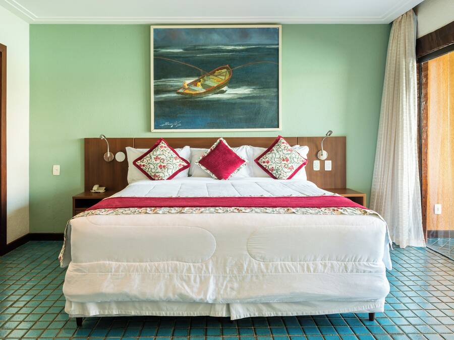 A hotel room with a large bed, a painting of a boat on the wall, and a view of the ocean through the window.