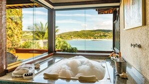 A bathroom with a large bathtub, a window with a view of a beach and hills, and a towel rack with towels. - Hotel Ferradura Private (Búzios)