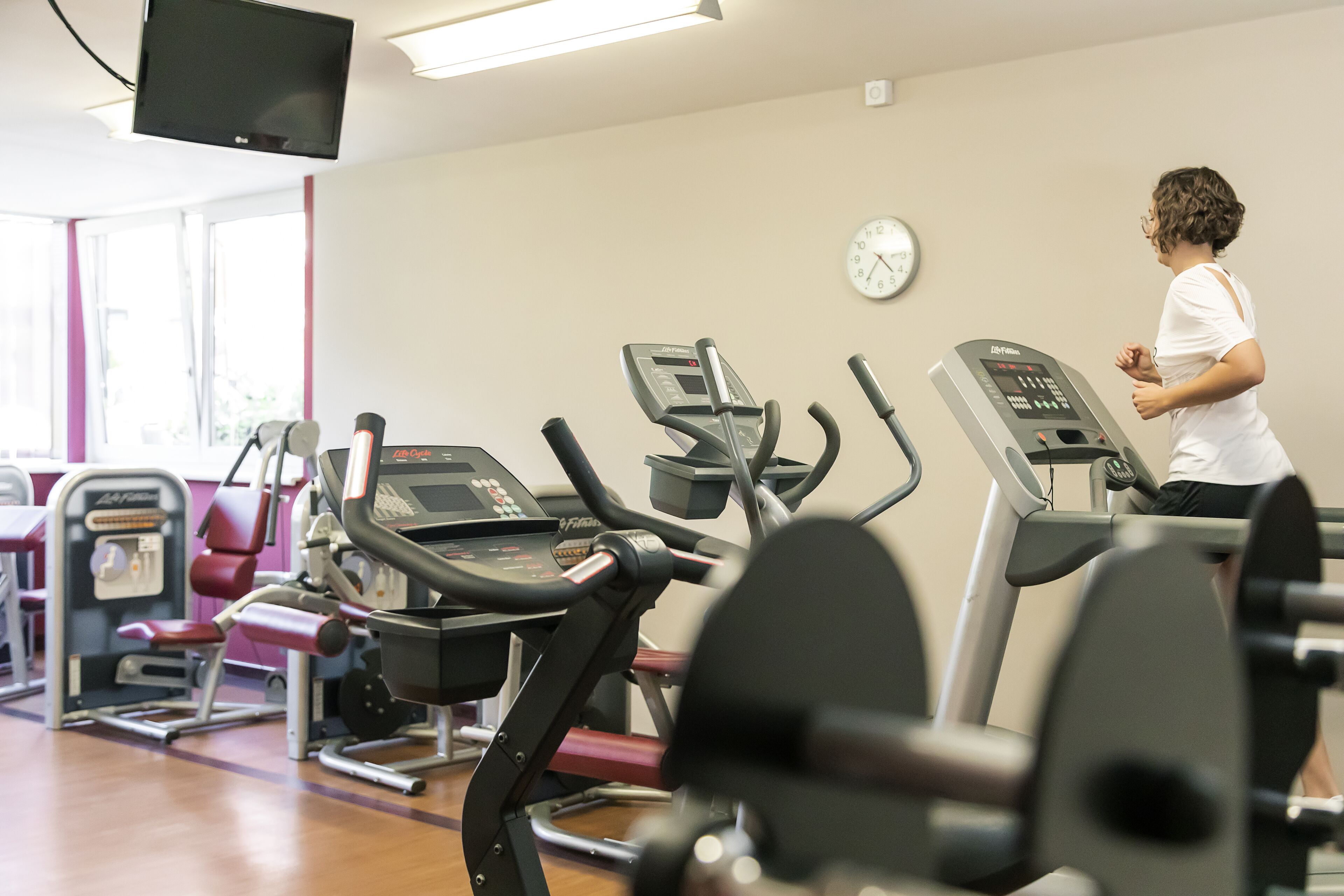 Un gimnasio con varias máquinas de ejercicio, un reloj en la pared y una persona usando una cinta de correr.
