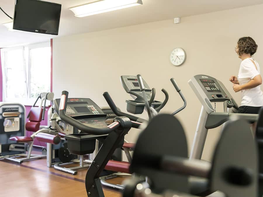 Un gimnasio con varias máquinas de ejercicio, un reloj en la pared y una persona usando una cinta de correr.