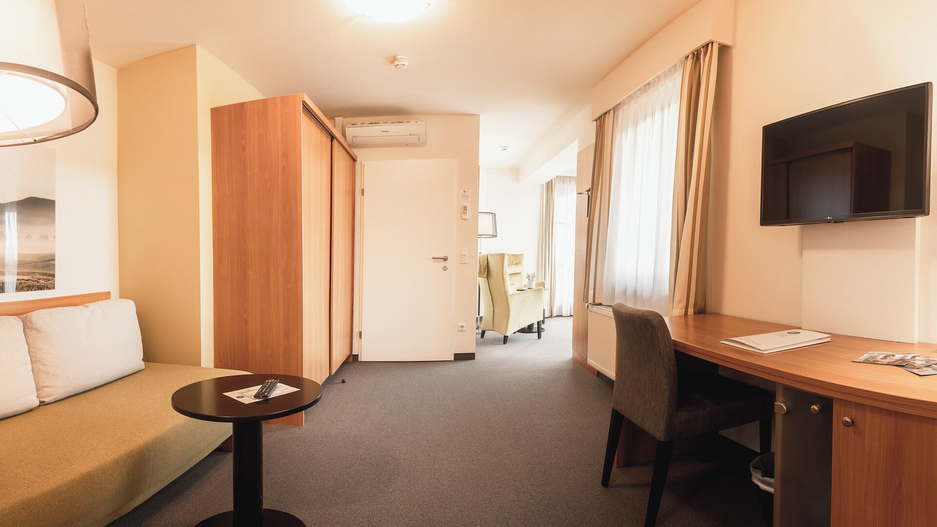 Habitación de hotel con sofá, escritorio, silla, televisor y ventana con cortinas.