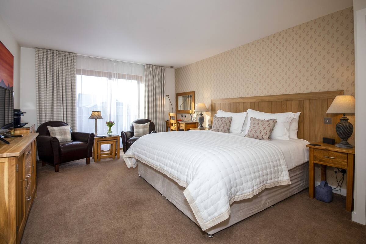A hotel room with a large bed, two chairs, a desk, a lamp, and a mirror.