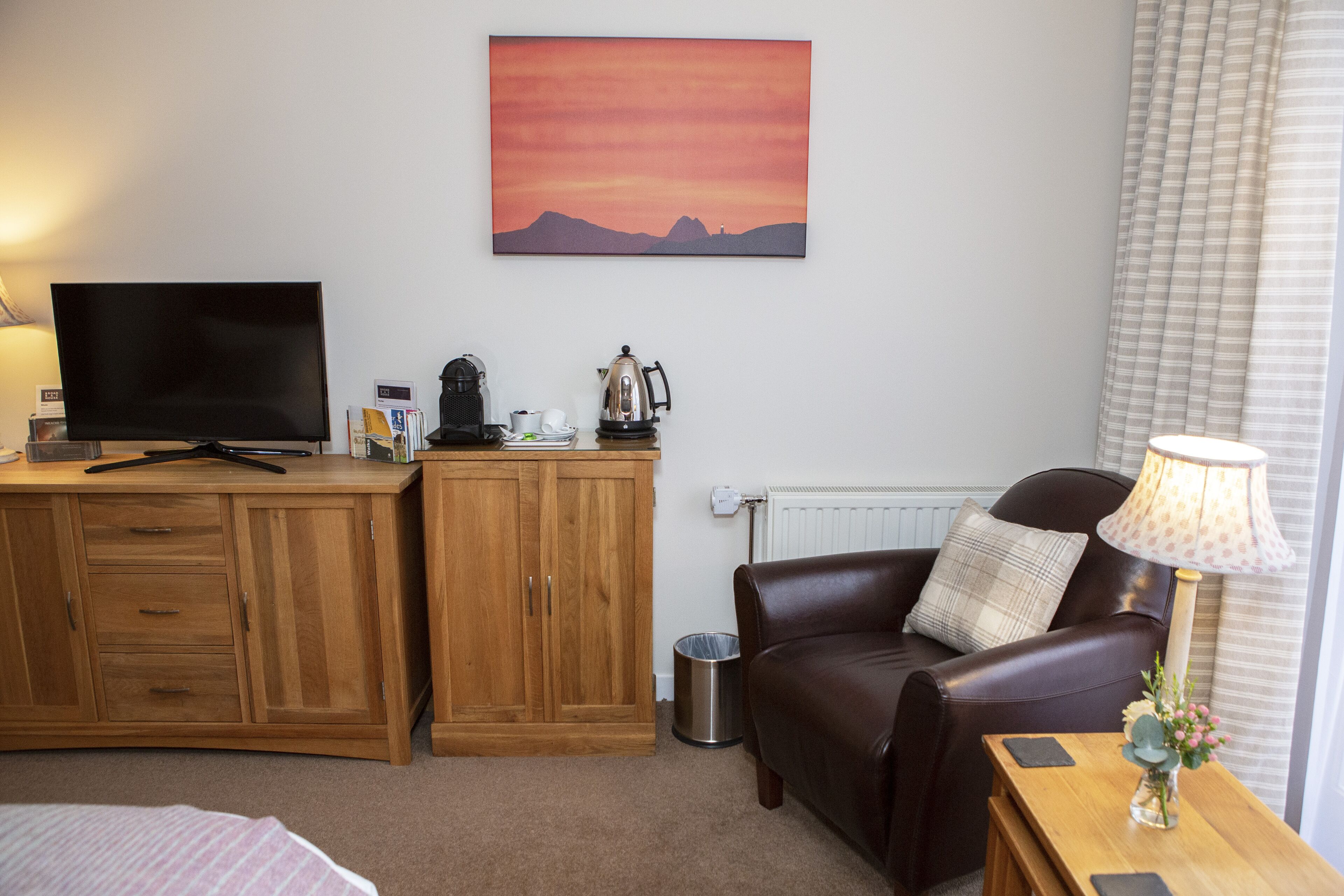 Ein gemütliches Zimmer mit einem Holztisch, einem Fernseher, einer Kaffeemaschine, einem Ledersofa und einem Sonnenuntergangsbild.