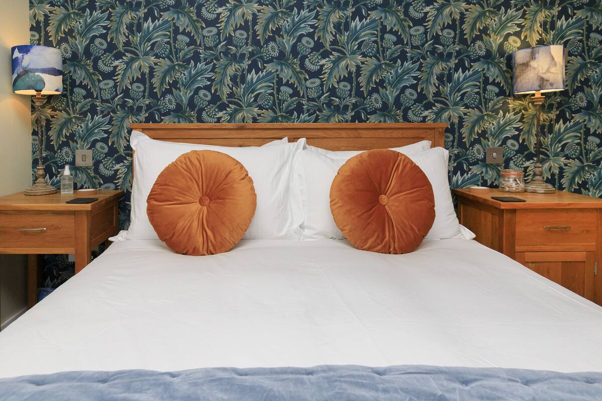 A neatly made bed with two large orange pillows, a wooden headboard, and a floral wallpaper pattern. There are two bedside tables with lamps and a small nightstand with a lamp.