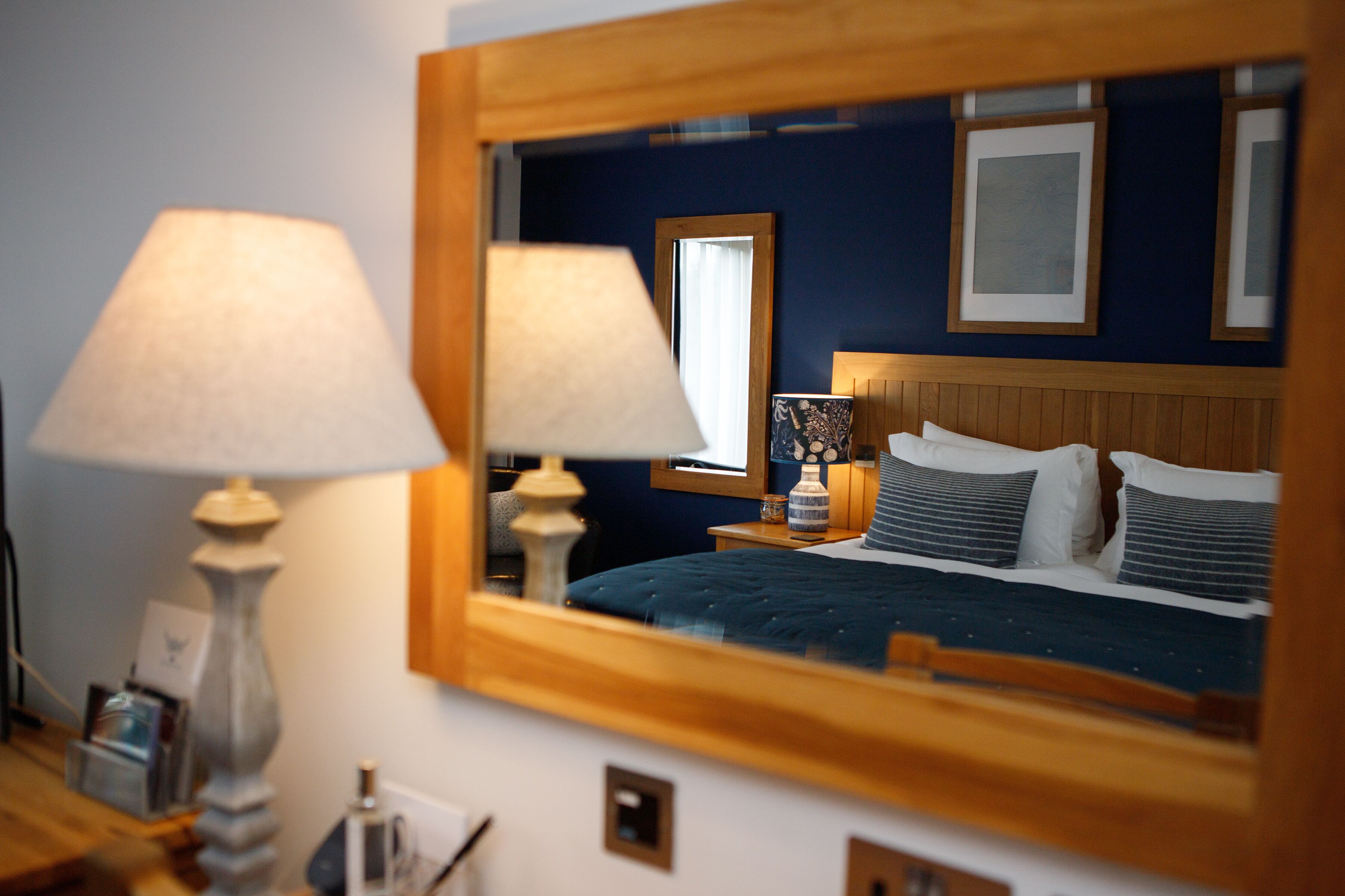 A hotel room with a large mirror reflecting the bed and nightstands, a lamp, and a framed picture on the wall.