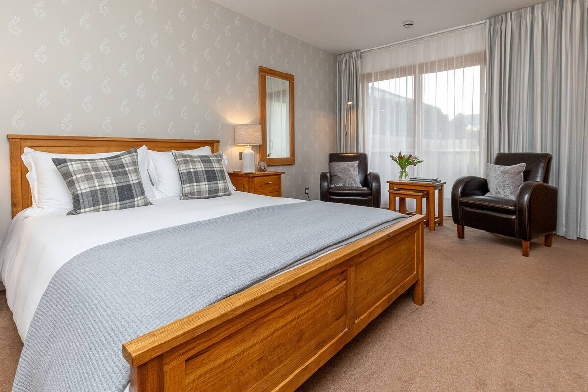 A hotel room with a large wooden bed, two chairs, a small table, a mirror, and a window with curtains.