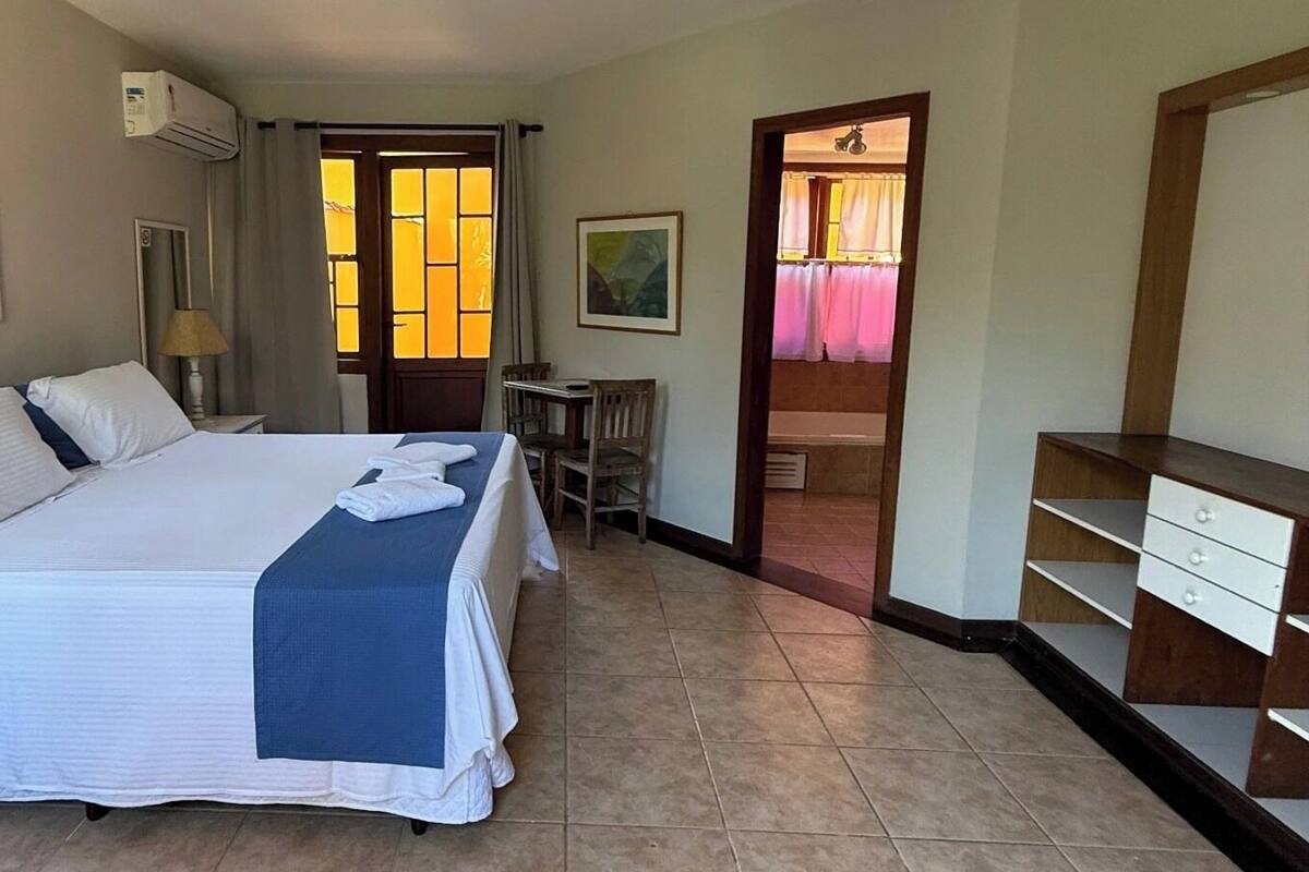 Habitación de hotel con una cama grande, un escritorio, una silla y una mesita. Se observa una puerta que da a otra habitación y una ventana con cortinas.