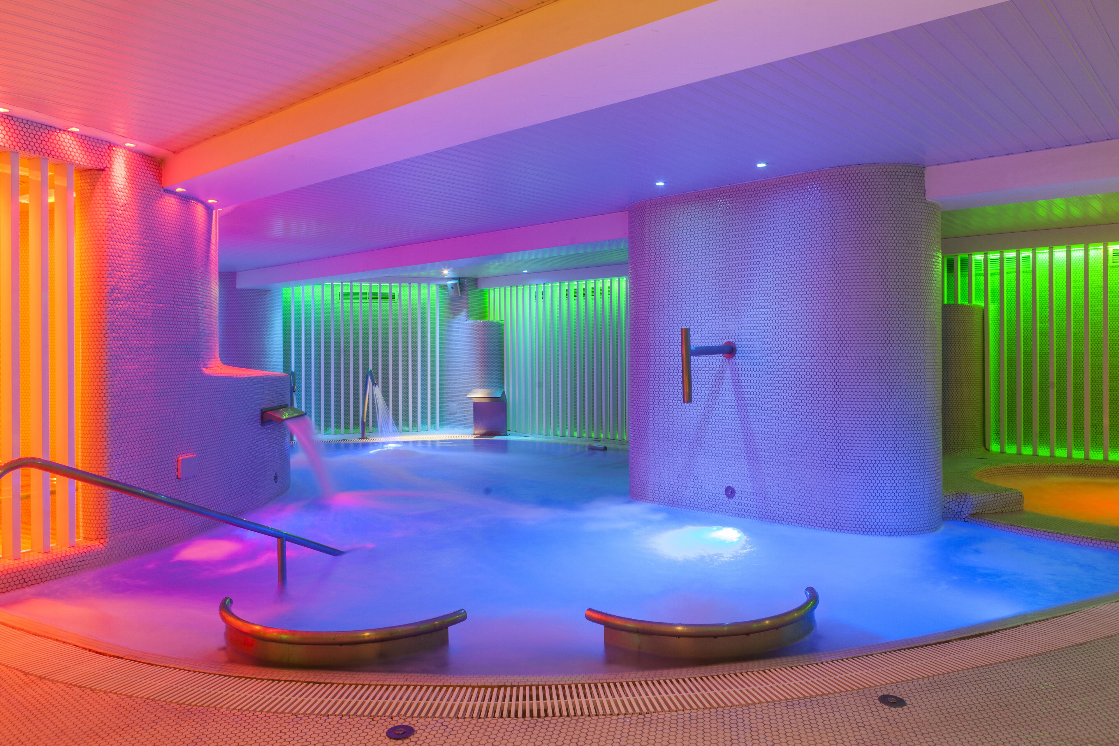 Un área de spa moderna con suelo calefaccionado, una zona de estar con bancos y una pared con paneles de luz verticales.