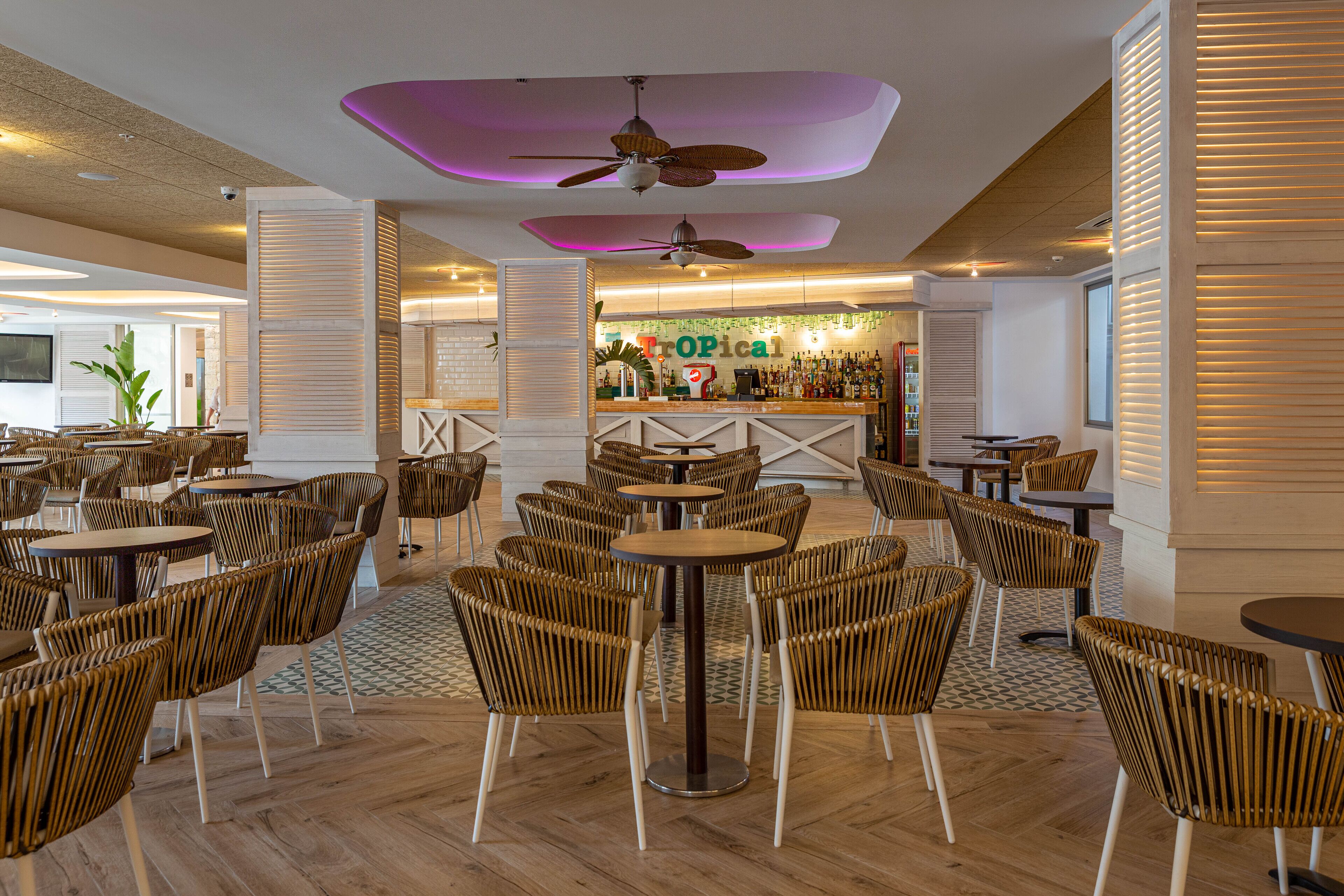 Un vaste espace intérieur doté de plusieurs tables rondes et de chaises en osier, un coin bar à l’ambiance tropicale et des ventilateurs de plafond.