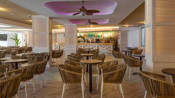 Un vaste espace intérieur doté de plusieurs tables rondes et de chaises en osier, un coin bar à l’ambiance tropicale et des ventilateurs de plafond.