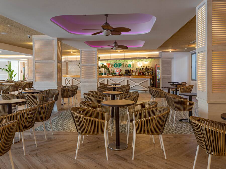 Un vaste espace intérieur doté de plusieurs tables rondes et de chaises en osier, un coin bar à l’ambiance tropicale et des ventilateurs de plafond.