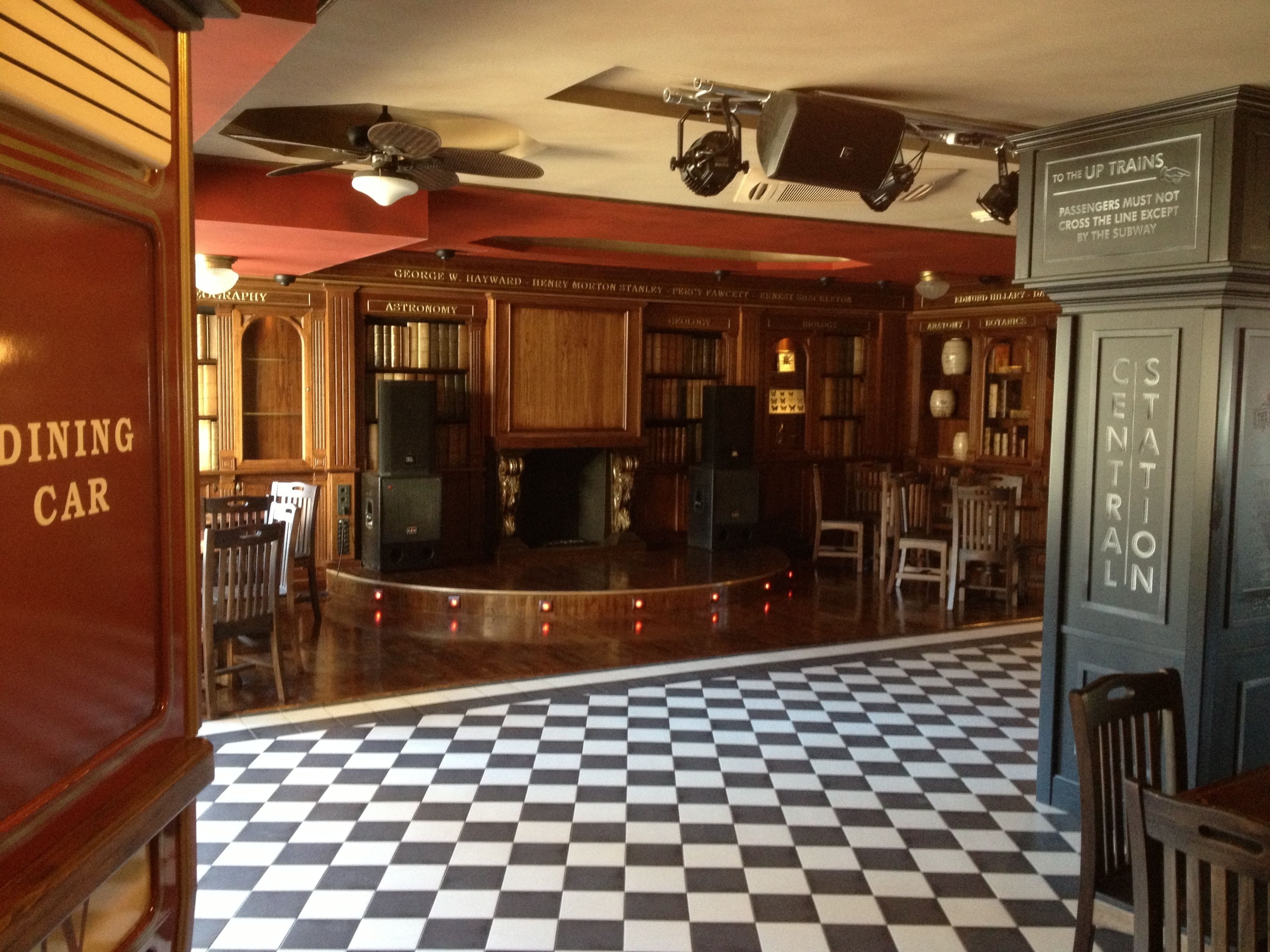 Interior de um antigo vagão-restaurante com piso xadrez, painéis de madeira e um balcão central.