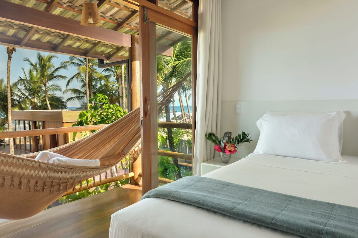 A bedroom with a bed, a hammock, and a view of palm trees and the ocean.