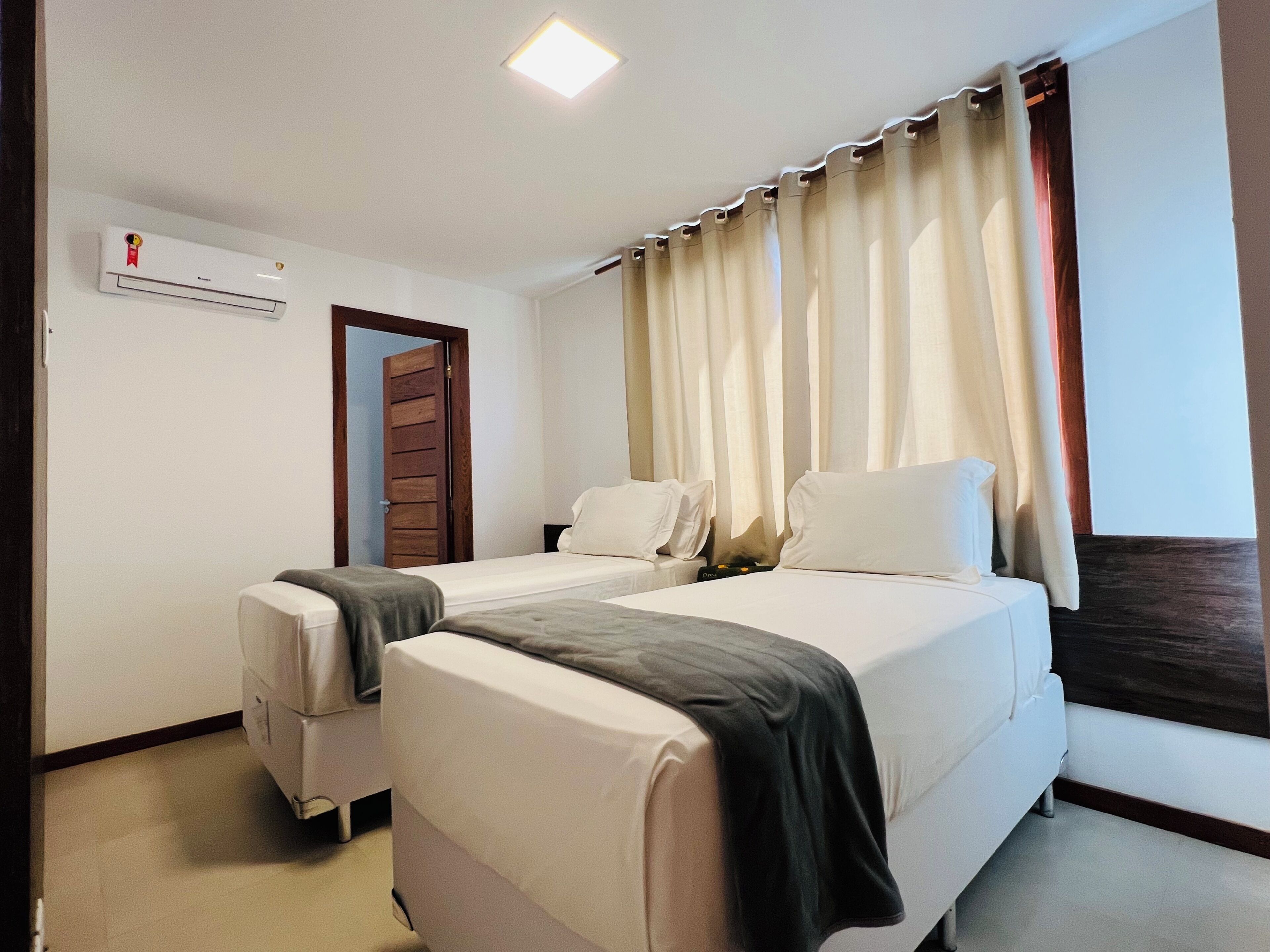 A hotel room with two beds, a ceiling light, an air conditioning unit, and a window with curtains.