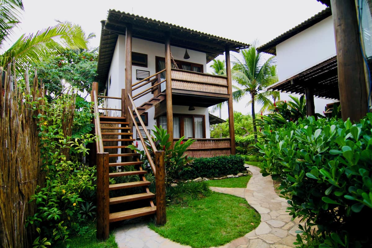 A wooden staircase leading to a white building with a balcony, surrounded by lush greenery and a stone pathway.