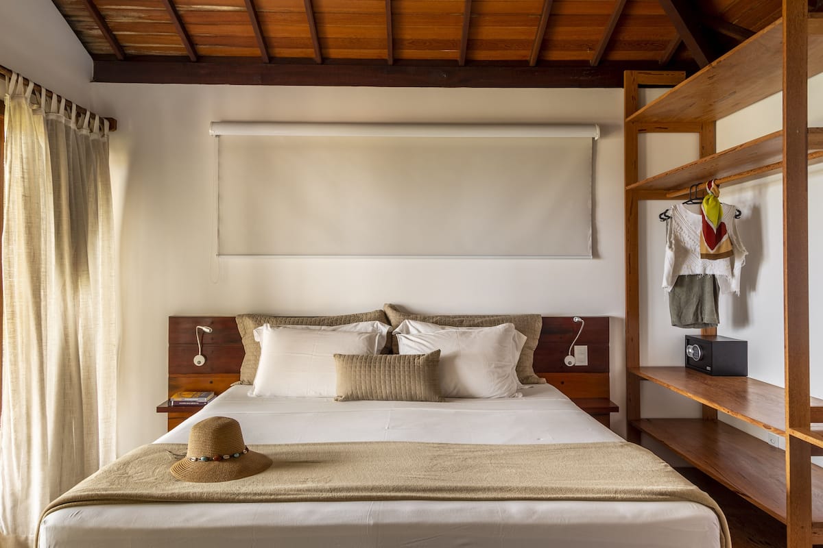A neatly made bed with a hat, a wooden headboard, a shelf with clothes, and a window with curtains.
