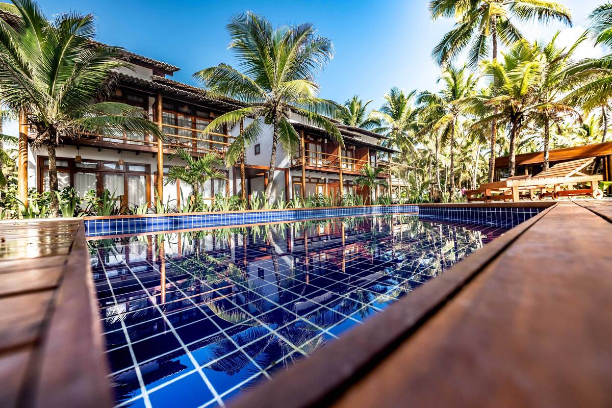 A pool with a tiled edge, surrounded by palm trees and a building with a balcony.