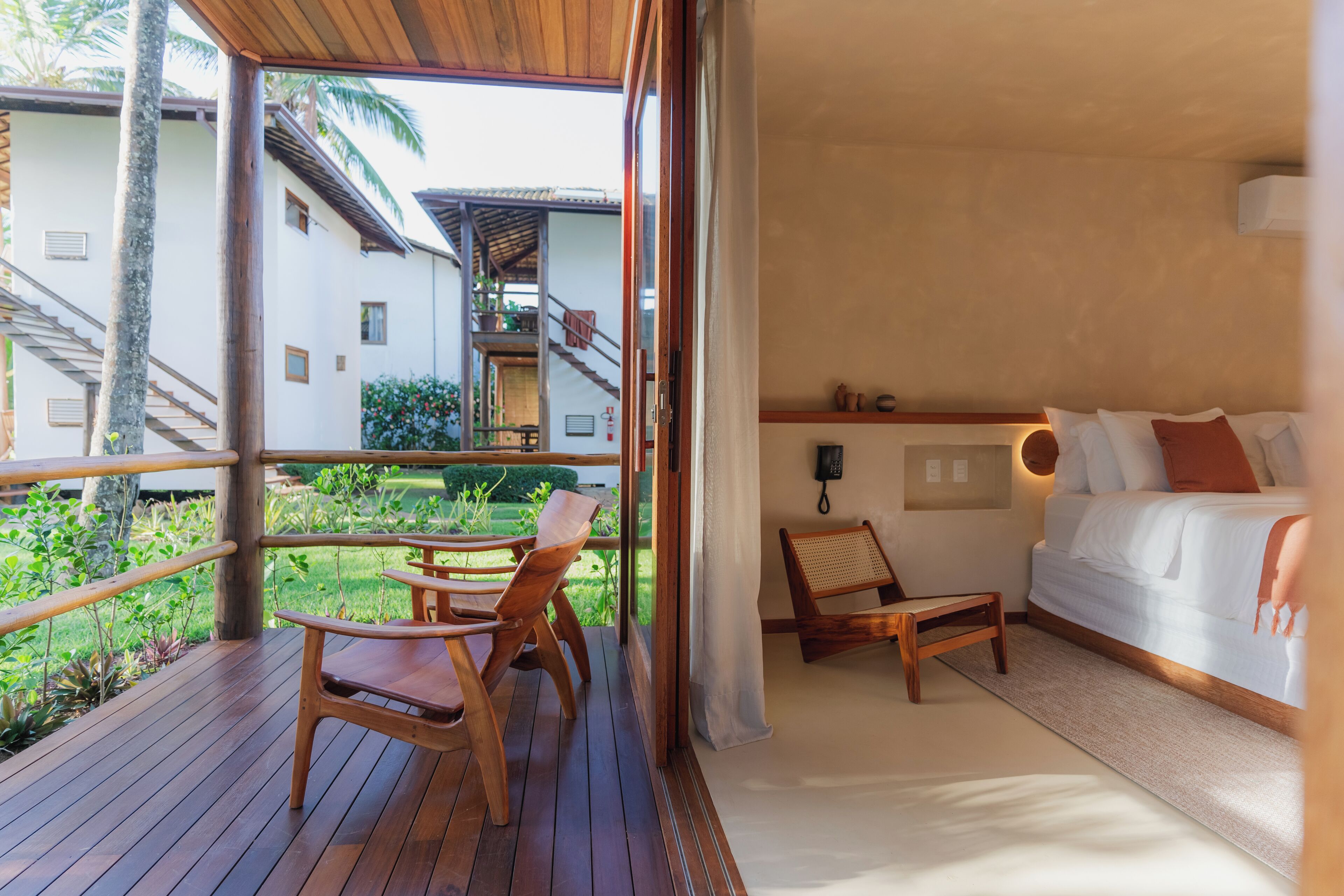 Een hotelkamer met balkon, houten meubilair, een bed en uitzicht op de tuin en andere gebouwen.