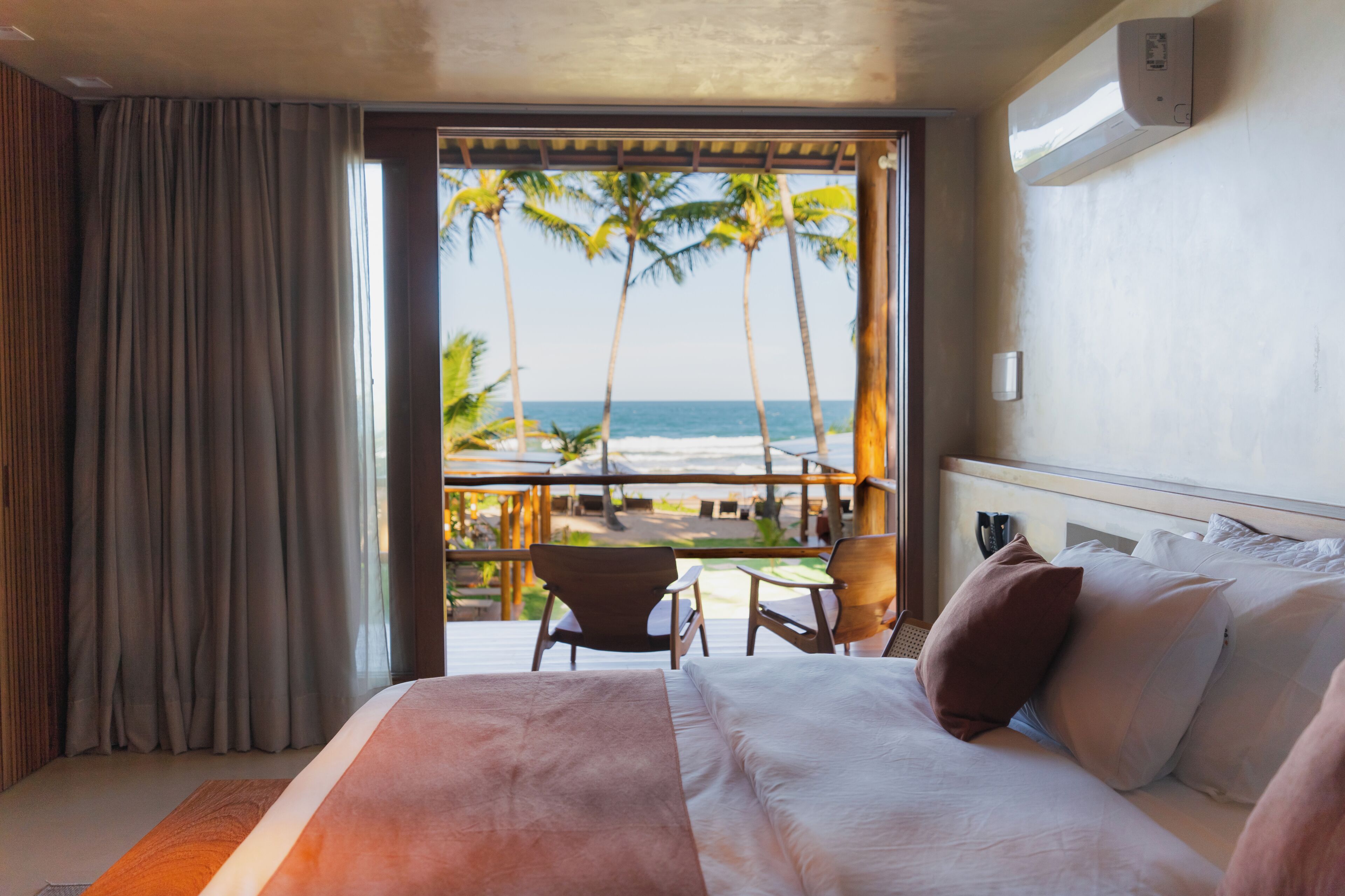 A hotel room with a bed, a balcony with a table and chairs, and a view of the beach and palm trees.