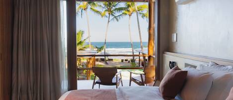 A hotel room with a bed, a balcony with a table and chairs, and a view of the beach and palm trees.