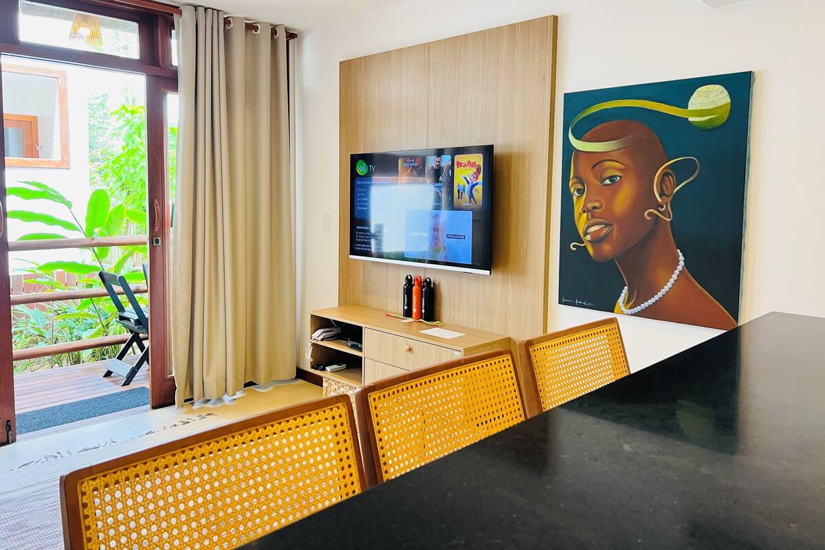 A modern hotel room with a flat-screen TV mounted on the wall, a dining area with yellow woven chairs, and a large painting of a woman.
