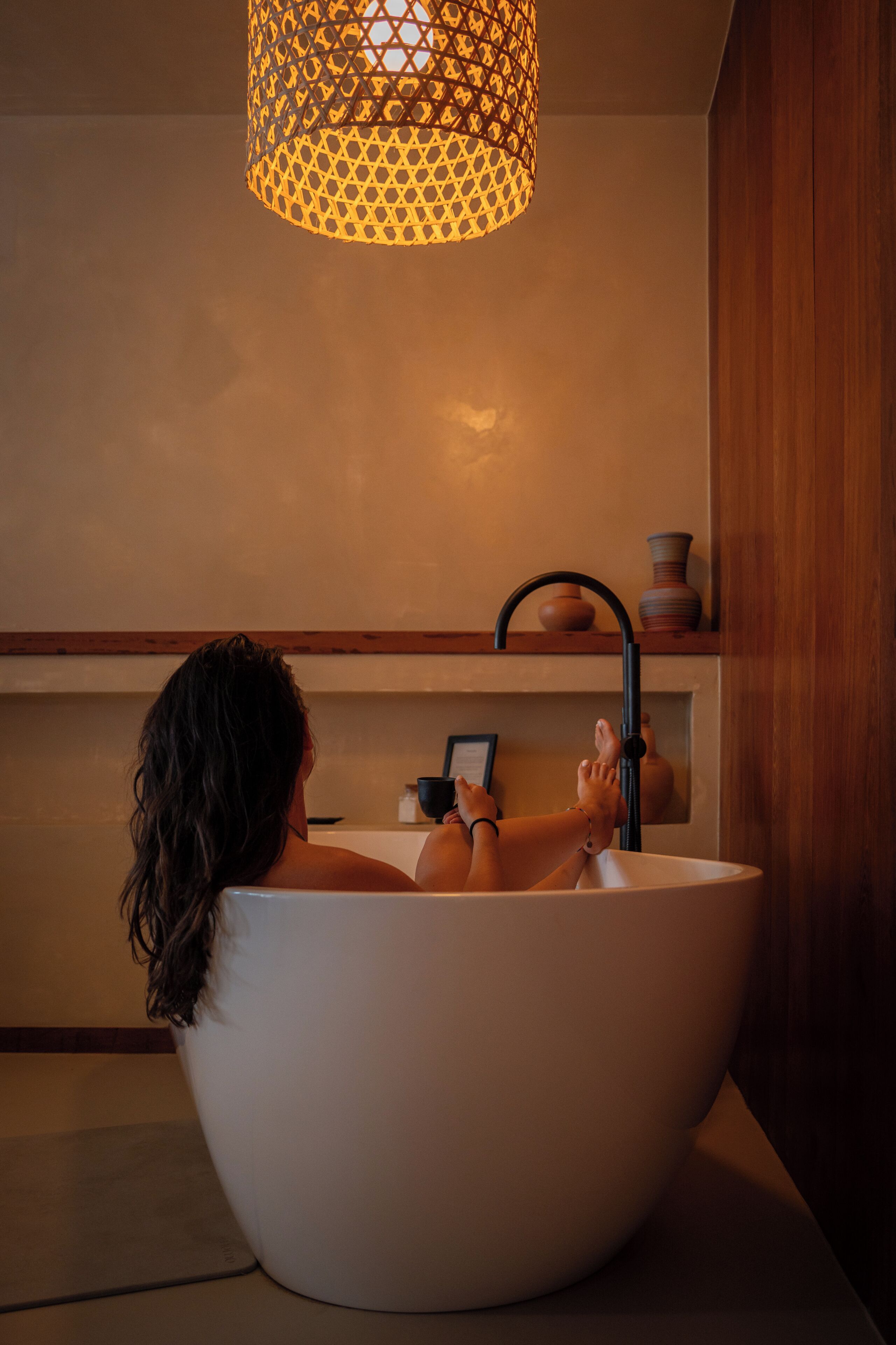 A person relaxing in a large white bathtub with a black faucet, a wooden shelf with decorative items, and a patterned hanging light fixture.