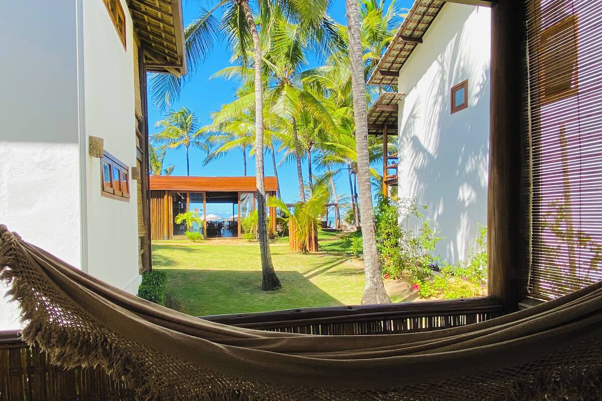 A view through a window with a woven blind, featuring a patio area with a wooden pergola, palm trees, and a white building with a small window.