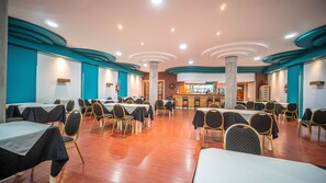Uma ampla área de refeições com várias mesas e cadeiras, um bar ao fundo e piso de madeira envernizada.