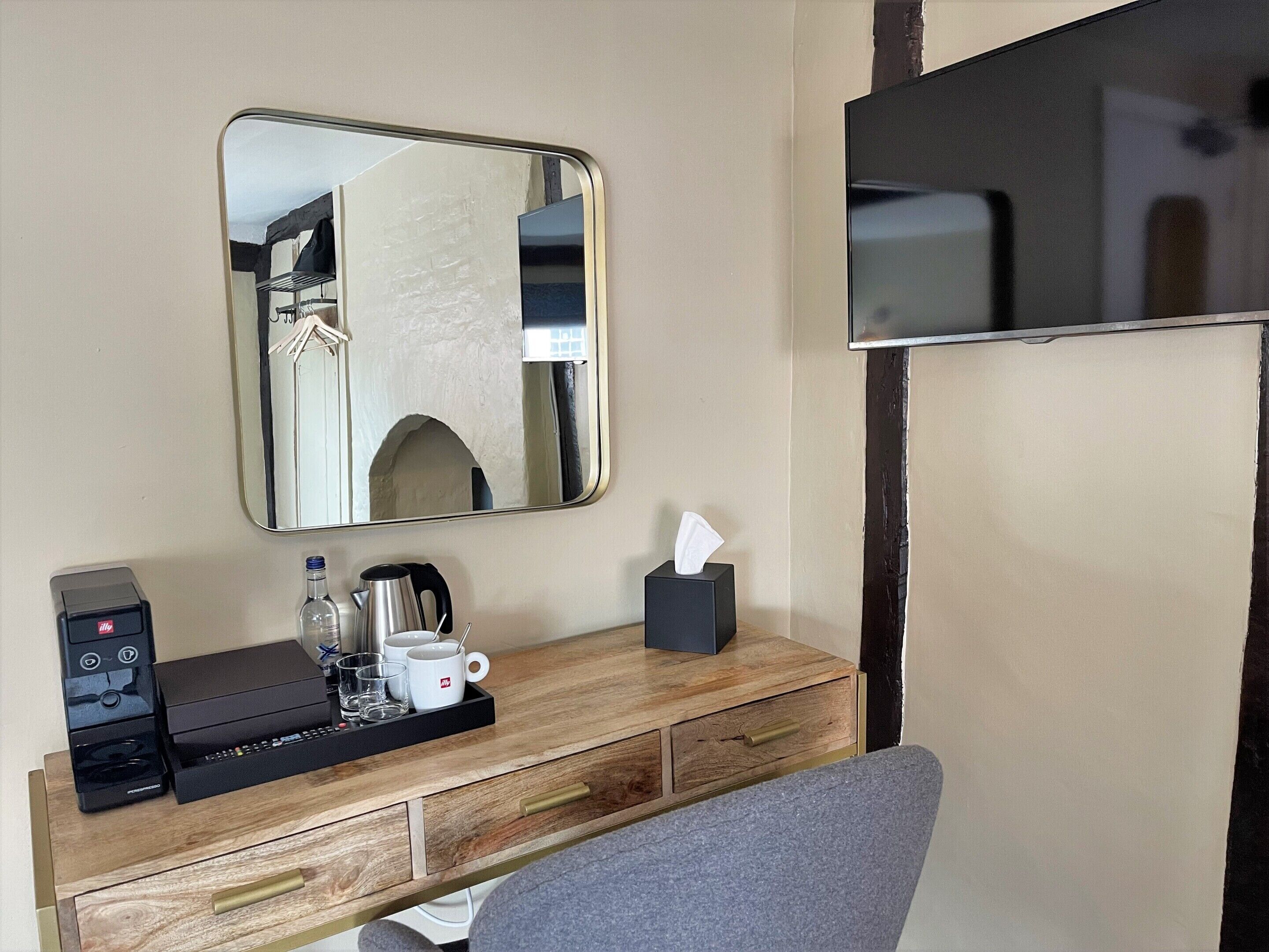 Ein Hotelzimmer mit einem Holztisch, einem Spiegel, einem Fernseher, einer Kaffeemaschine und einer Taschentuchbox.