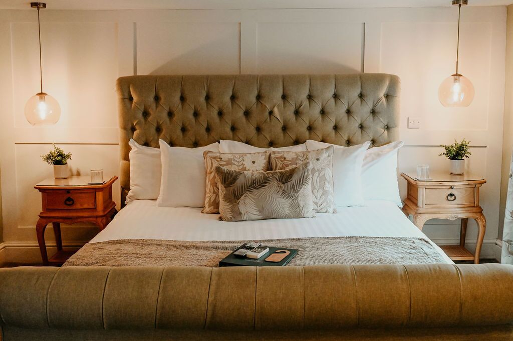Un dormitorio con cabecera capitonada, cama con ropa de cama blanca, dos mesitas de noche con lámparas y una planta en la mesita.