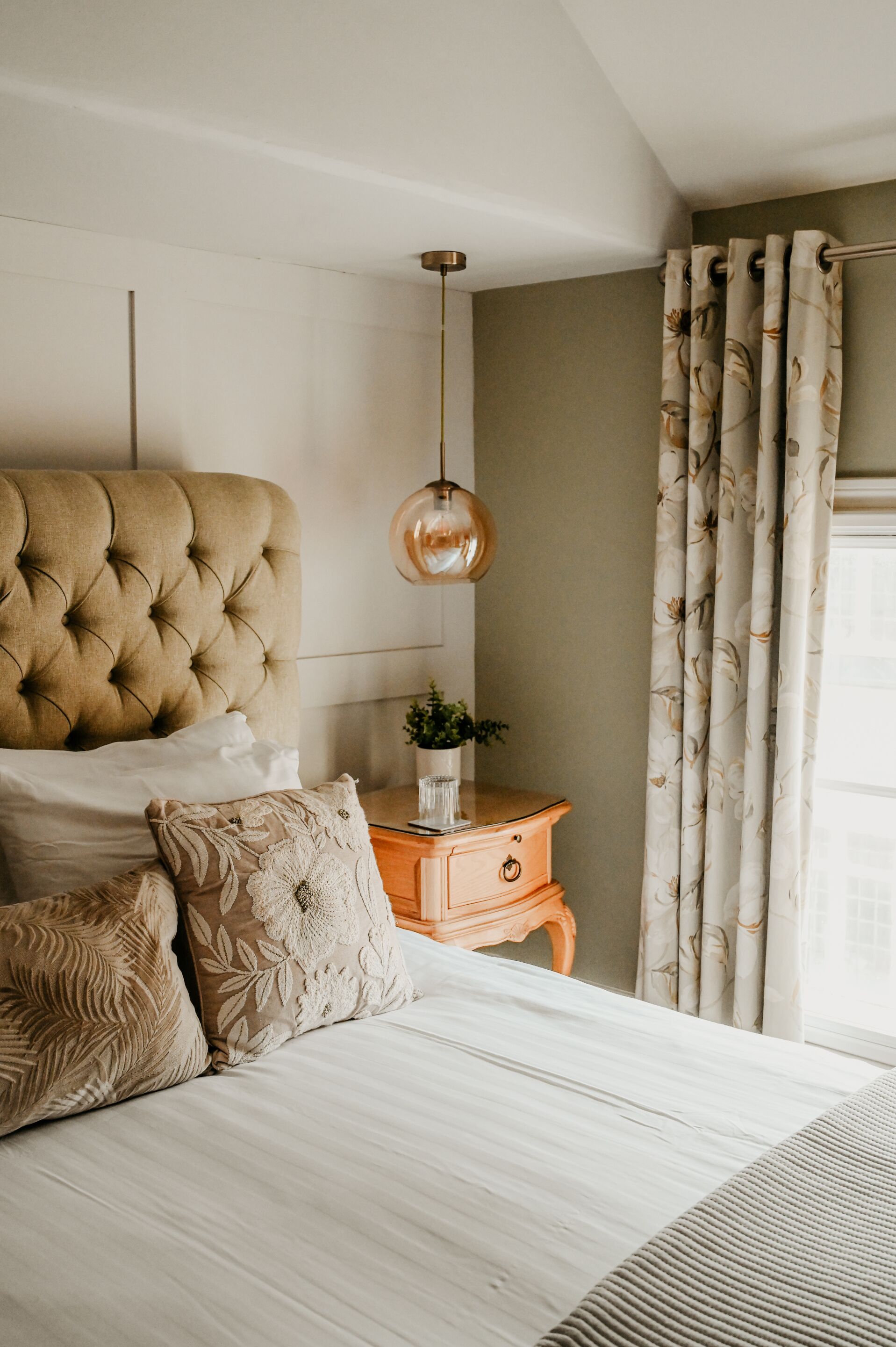 A bedroom with a tufted headboard, a bedside table with a plant, and a hanging pendant light.