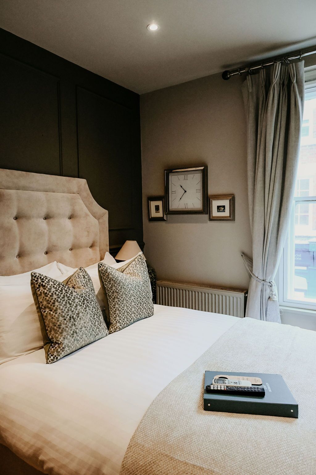 A hotel room with a tufted headboard, a bed with patterned pillows, a wall clock, a framed picture, and a window with curtains.