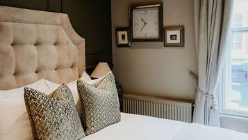 A hotel room with a tufted headboard, a bed with patterned pillows, a wall clock, a framed picture, and a window with curtains.