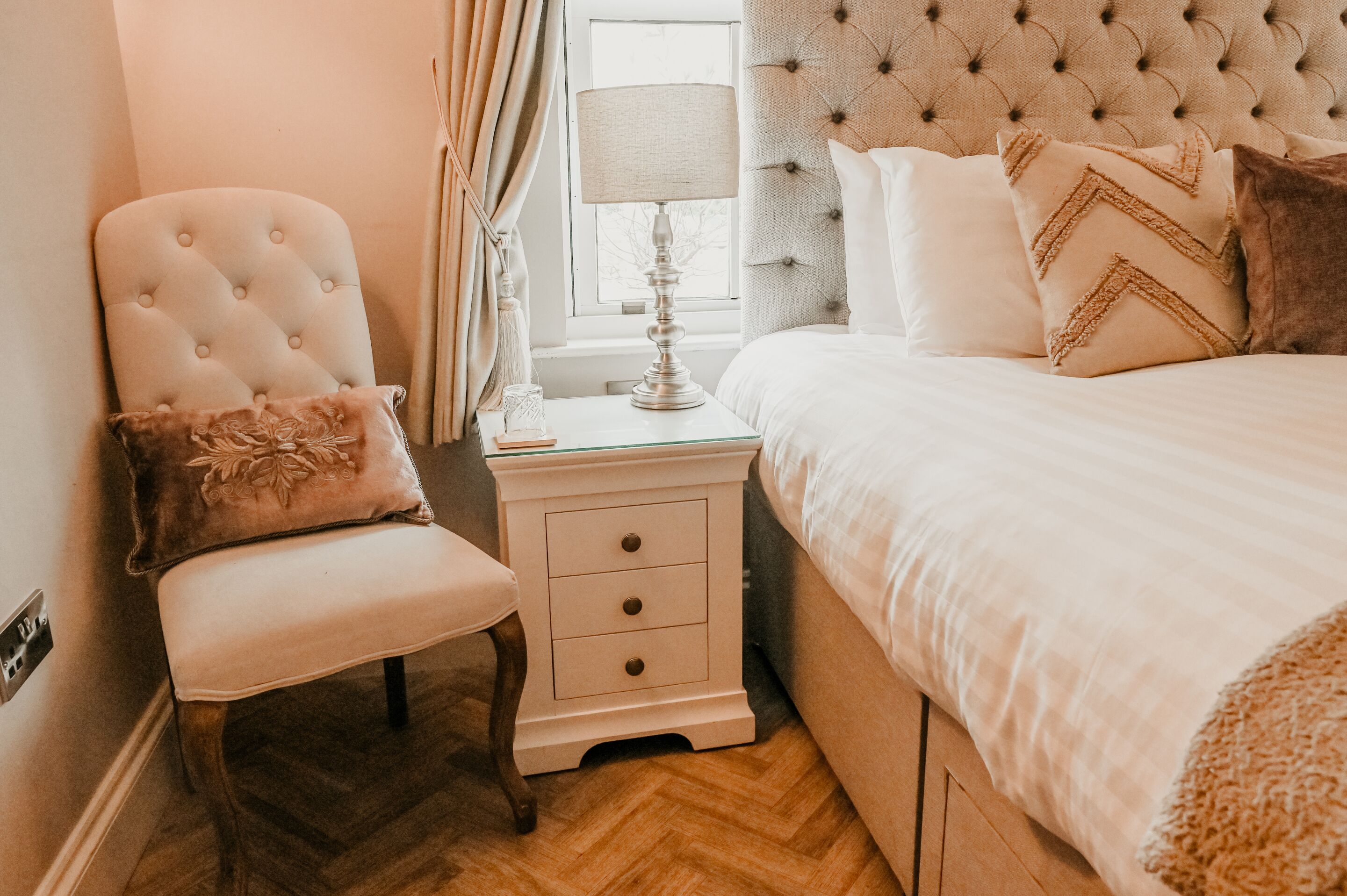 A bedroom with a tufted headboard, a bedside table with a lamp, a chair with a cushion, and a bed with pillows.