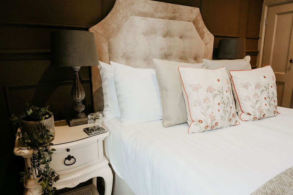 Un lit avec une literie blanche et des coussins à motifs floraux, une lampe de chevet, une petite table avec une plante et un panneau décoratif mural.