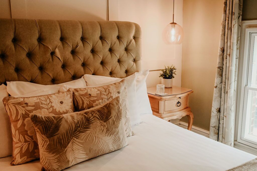 Una cama con cabecera capitonada, una mesita de noche con una planta y una ventana con cortinas.
