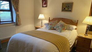 Una cama bien hecha con una colcha color crema y almohadones, un cabecero de madera, una lámpara de noche y una mesita de noche de madera con un cajón.