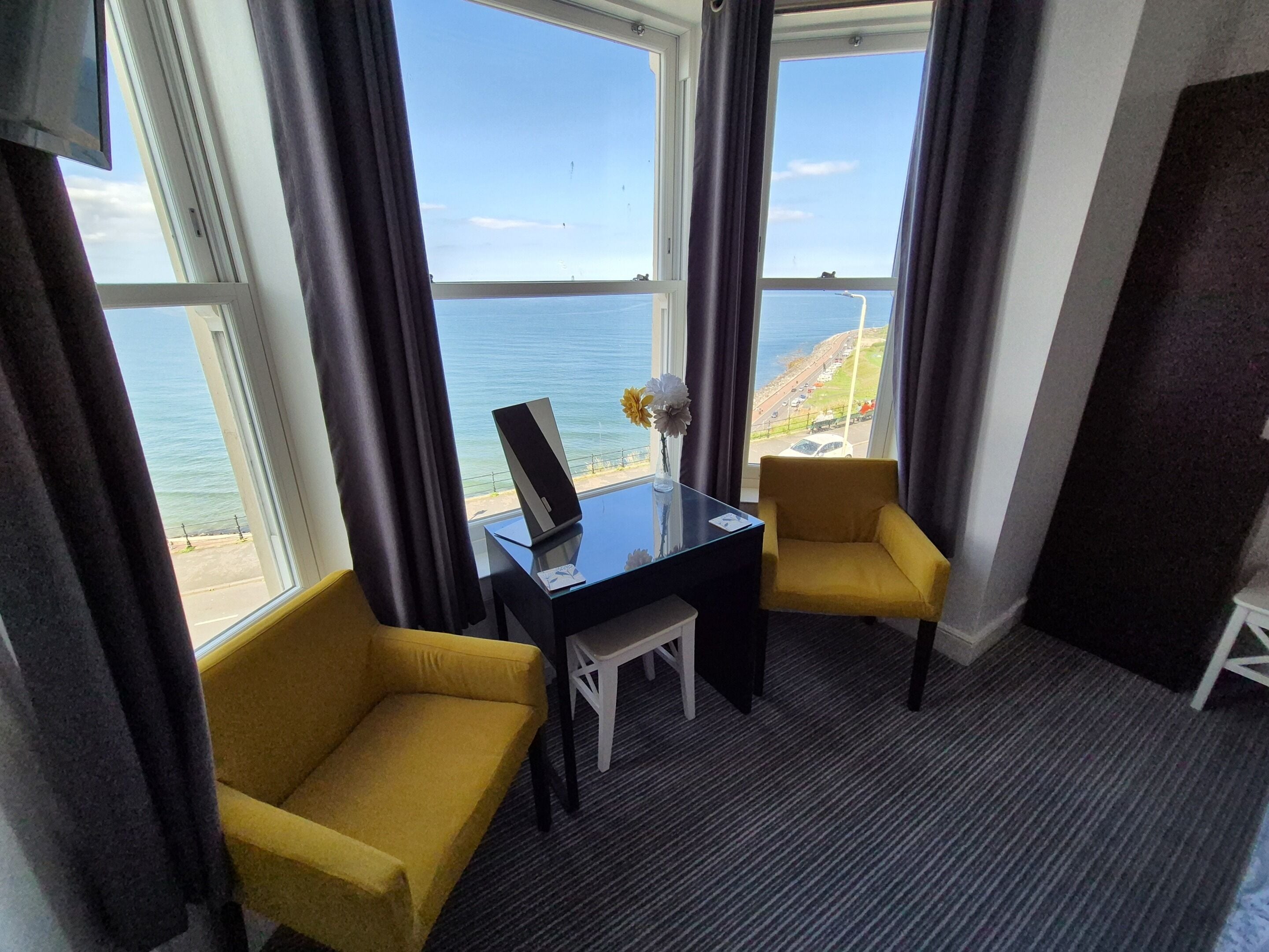 A hotel room with a desk, two yellow chairs, a small table, and a view of the sea.
