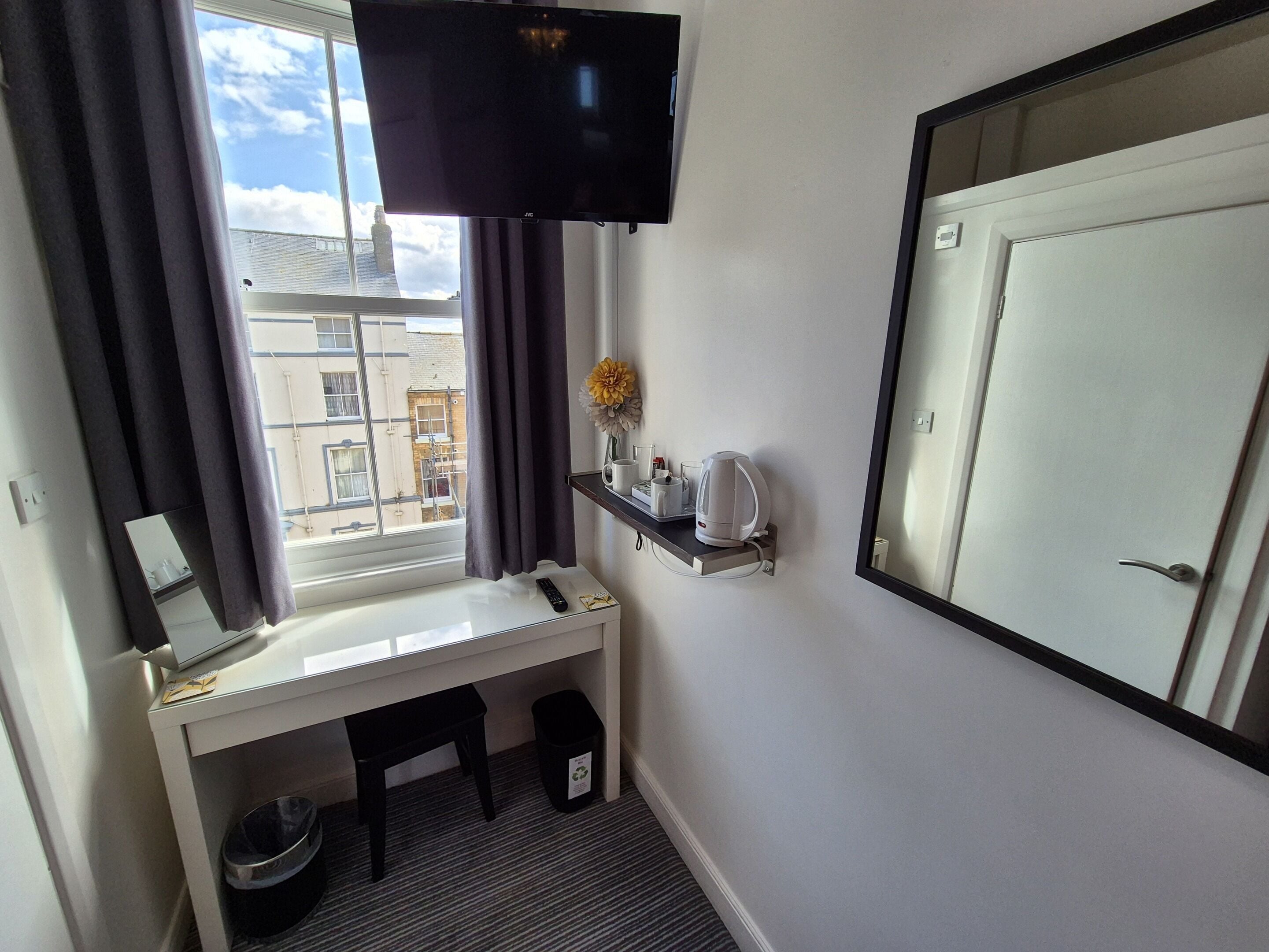 Een hotelkamer met een aan de muur bevestigde televisie, een bureau met een afstandsbediening, een koffiezetapparaat en een kleine prullenbak.