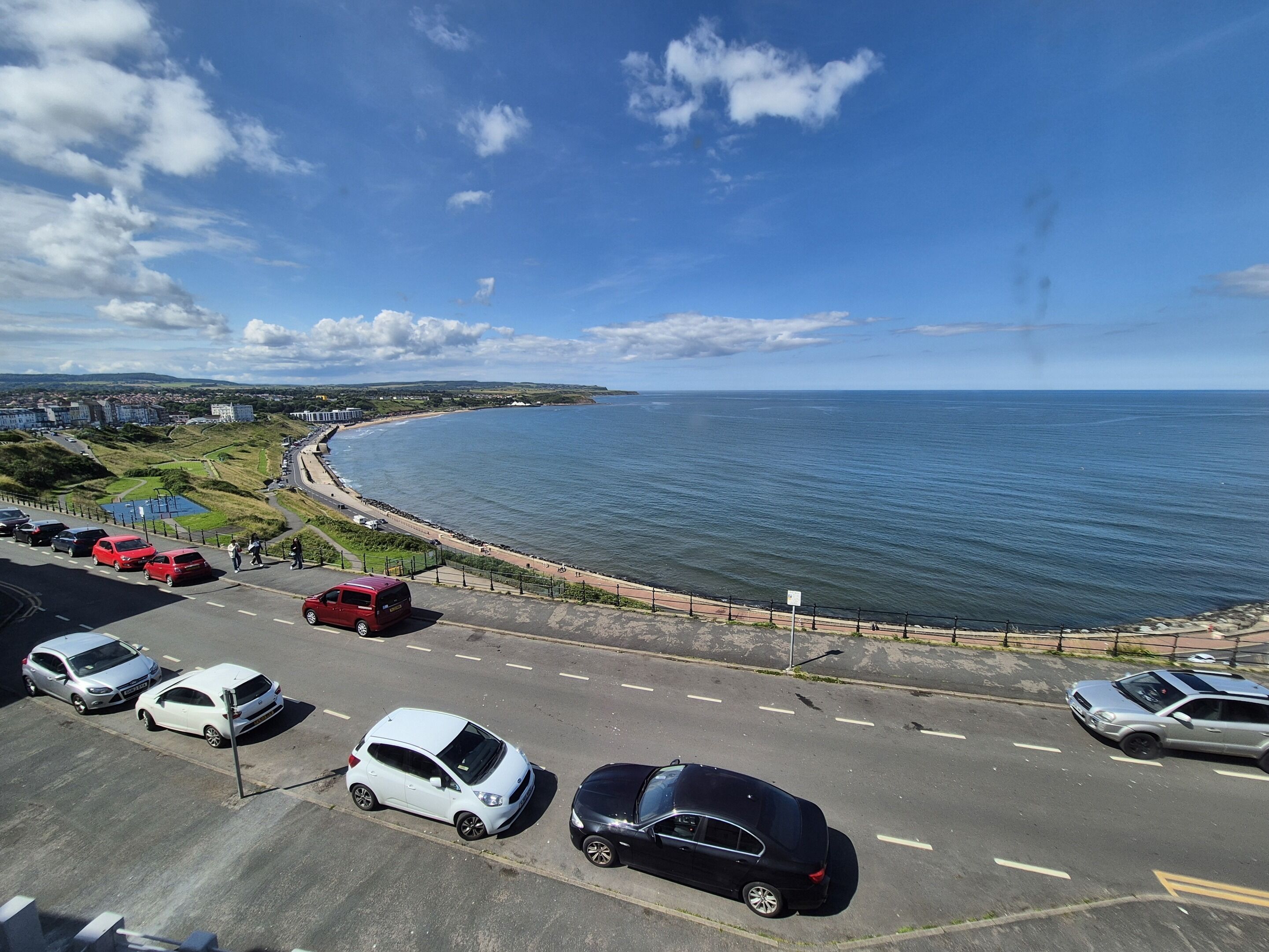 A coastal road with parked cars and a beach in the background.