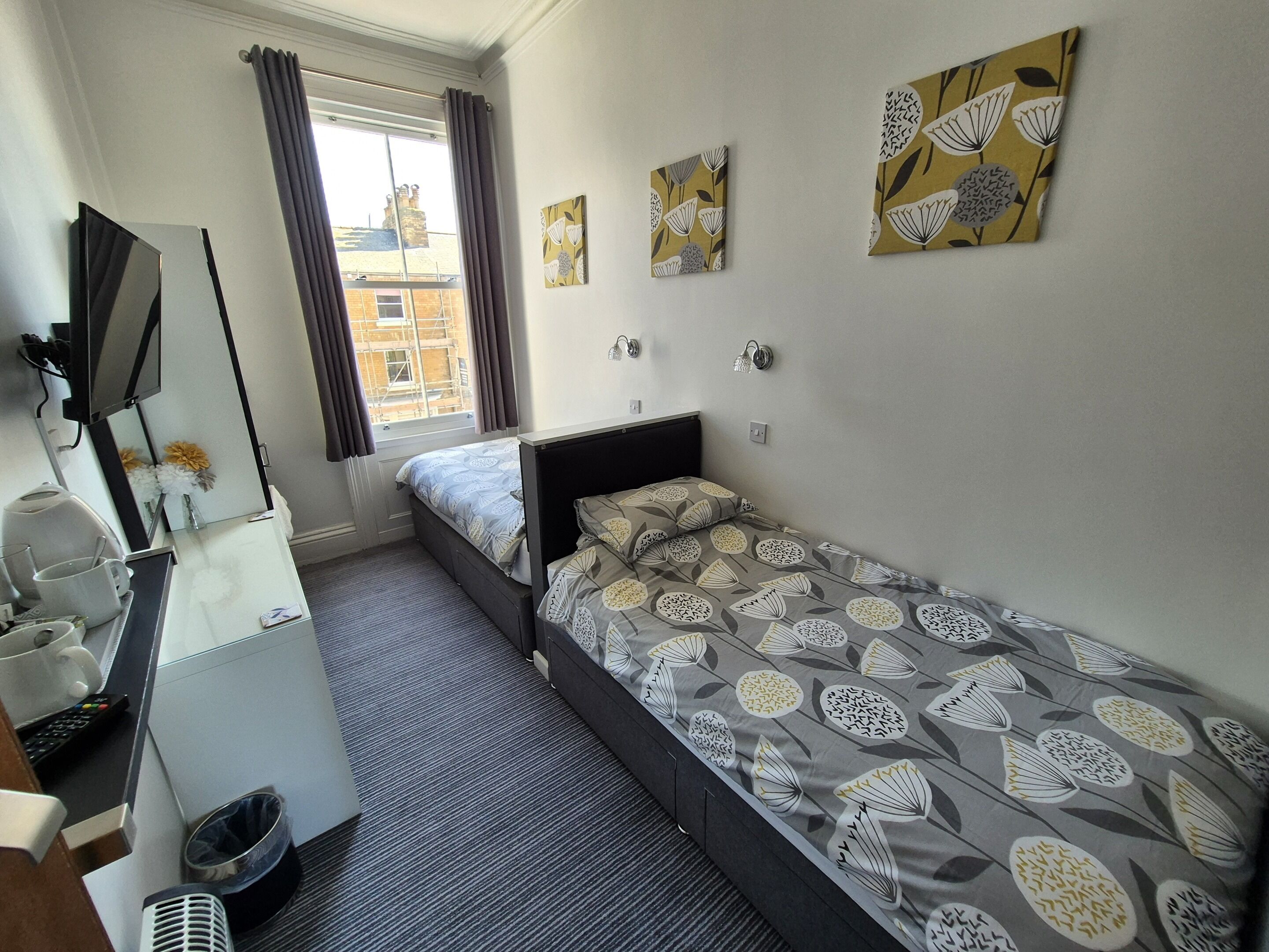 A compact hotel room with a single bed, a desk with a mirror, a small window with curtains, and two wall-mounted light fixtures.