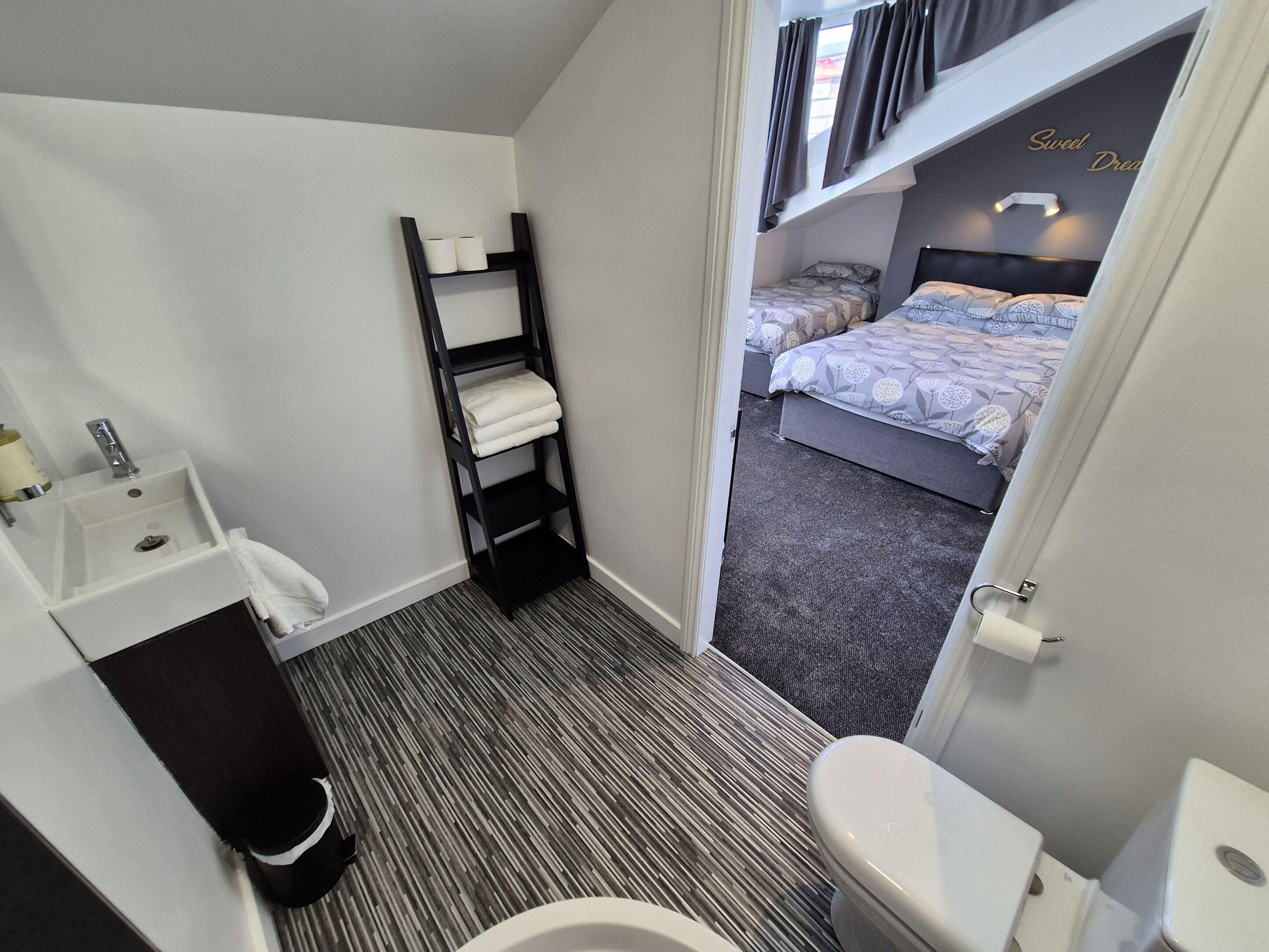 Habitación de hotel pequeña con baño, un lavamanos, un inodoro, un estante con toallas y una cama con ropa de cama estampada.