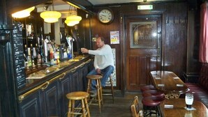 A bar area with wooden stools, a bar counter with various bottles, and a clock on the wall. - Nelson and Railway Inn (Nottingham)